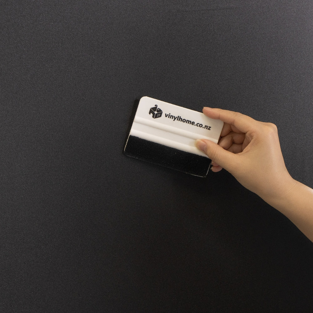 Hand holding a white squeegee with 'vinylhome.co.nz' branding against a blackout window film.