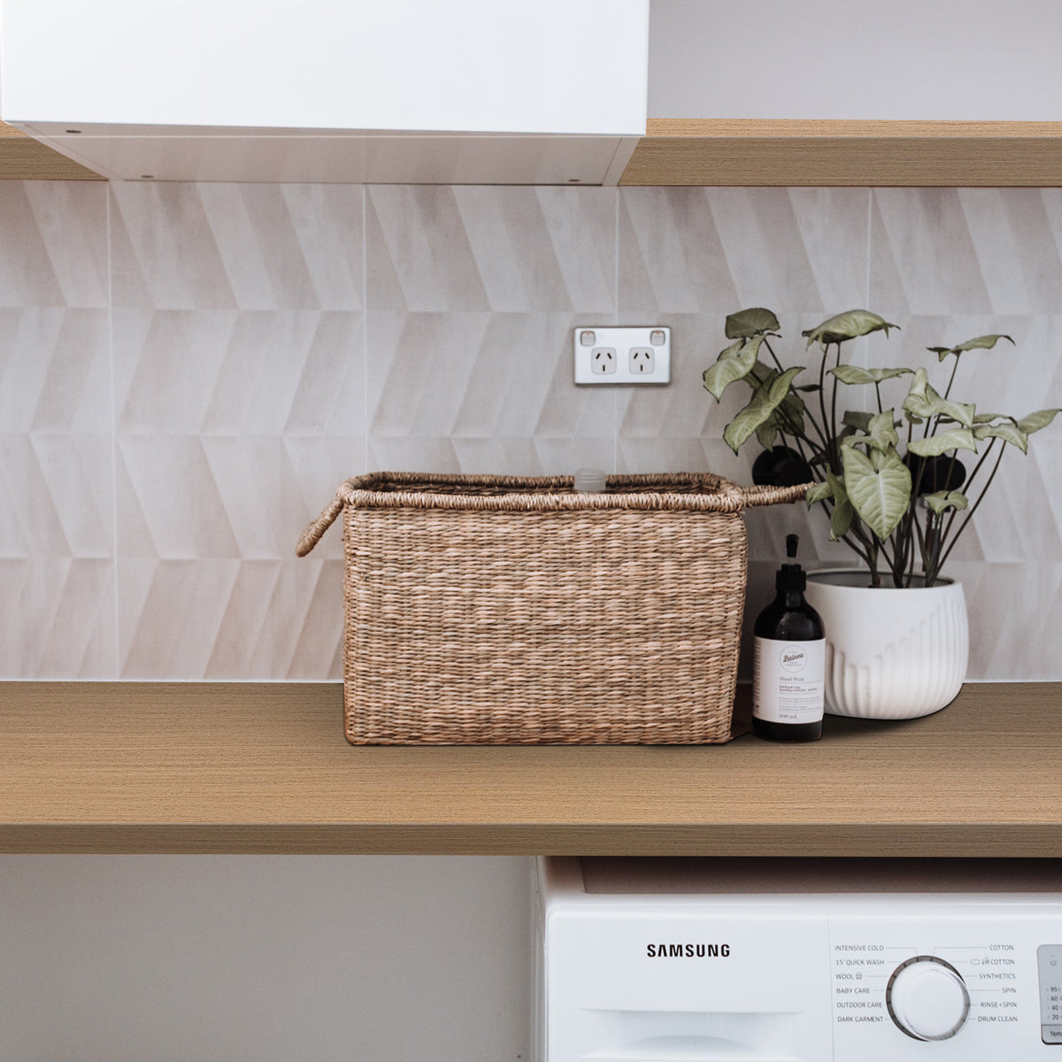 laundry benchtop with a woven basket, plant, and bottle against a geometric-patterned wall.