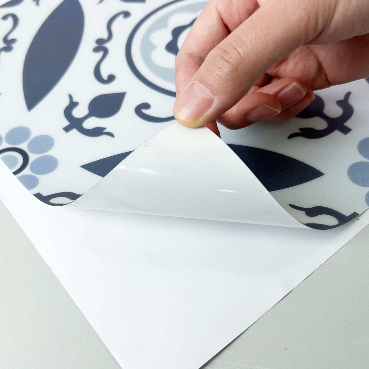 Hand peeling back a protective layer on a decorative tile with blue and white pattern.