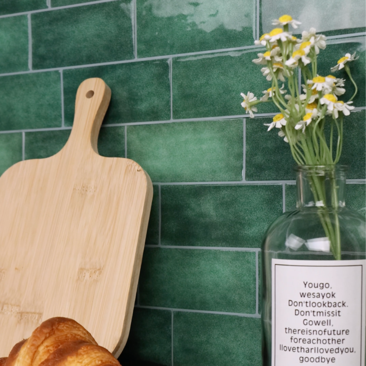 Wooden cutting board with a croissant, vase with flowers, and green tiled wall.