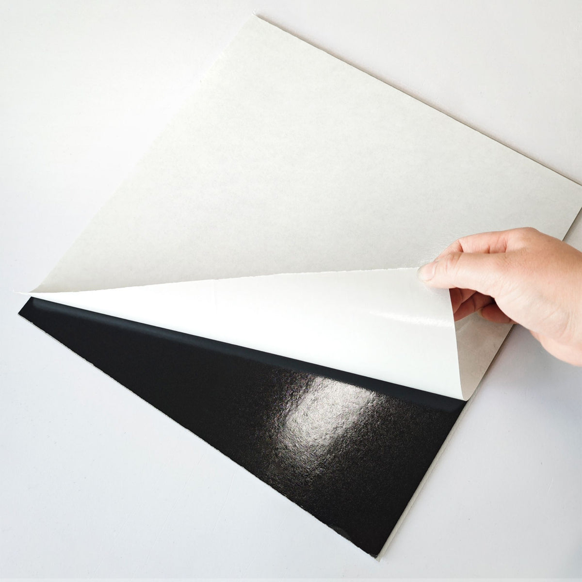 A hand peeling back a white backing sheet of a peel and stick tile, shown on a clean white background.