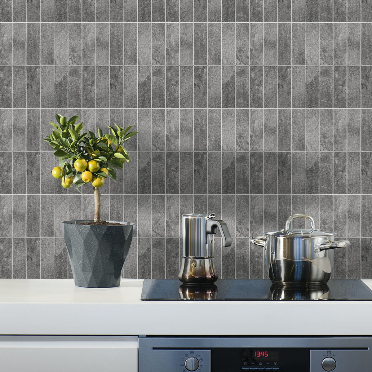 Grey rectangular stacked subway tile backsplash on a kitchen wall behind a white countertop with a black induction cooktop, a stainless steel pot, a coffee maker, and a potted lemon tree.