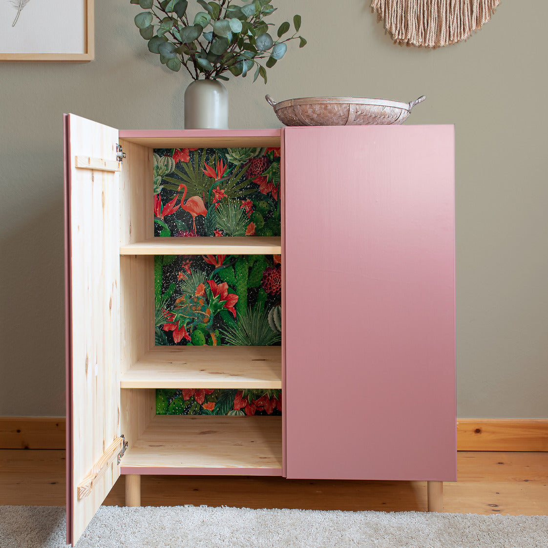 A pink wooden cabinet with one door open, showing light wood shelves and floral-patterned interior panels. The cabinet stands on a light rug against a beige wall with framed wall art and a plant on top.
