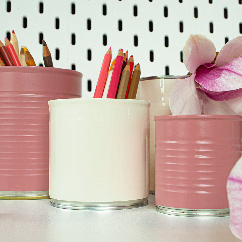Three ribbed metal can-style containers in dusty pink and off-white, arranged on a white surface against a white pegboard background. The pink containers hold colored pencils, and a pink orchid flower is placed in the right container.