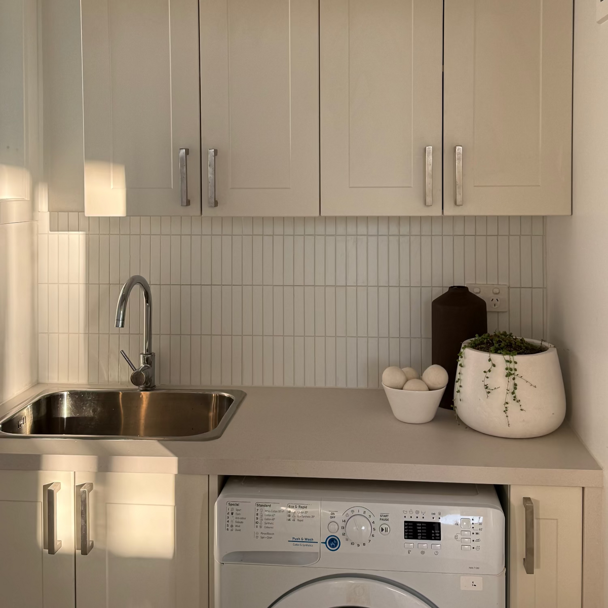 A bright, modern laundry or utility room corner with cream-colored cabinets, a stainless steel sink, a white tiled backsplash, and a white washing machine underneath. A dried floral arrangement sits on top of the upper cabinets, and a white planter with a trailing green plant is on the counter.