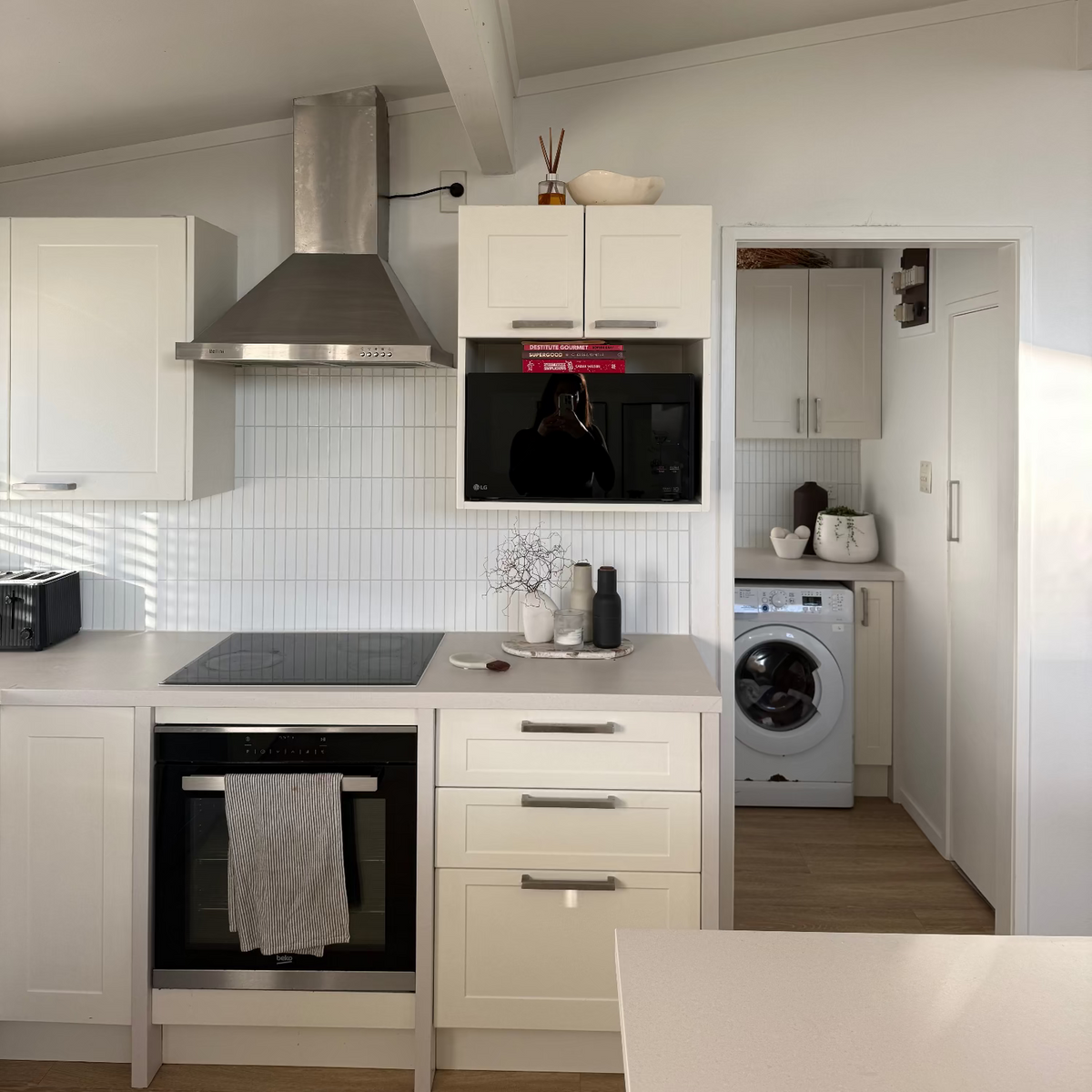 A modern kitchen interior with white cabinets, a stainless steel range hood, built-in black appliances, a tiled backsplash, and a washing machine visible in an adjacent room.