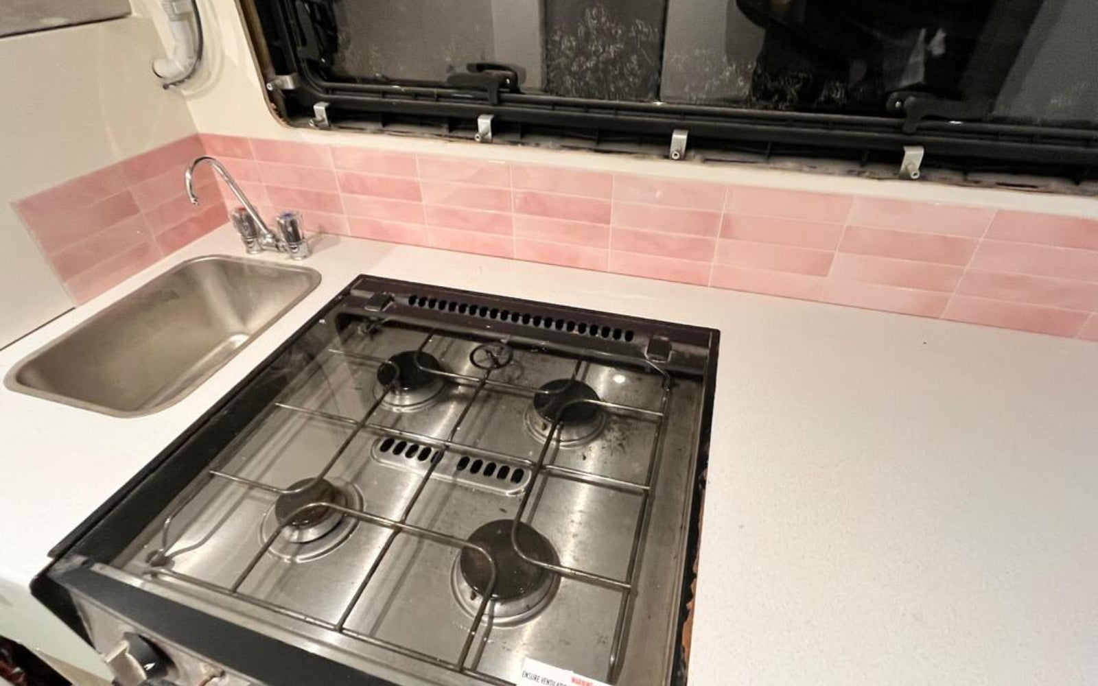 Stainless steel gas stove in a compact kitchen with pink tiled wall.