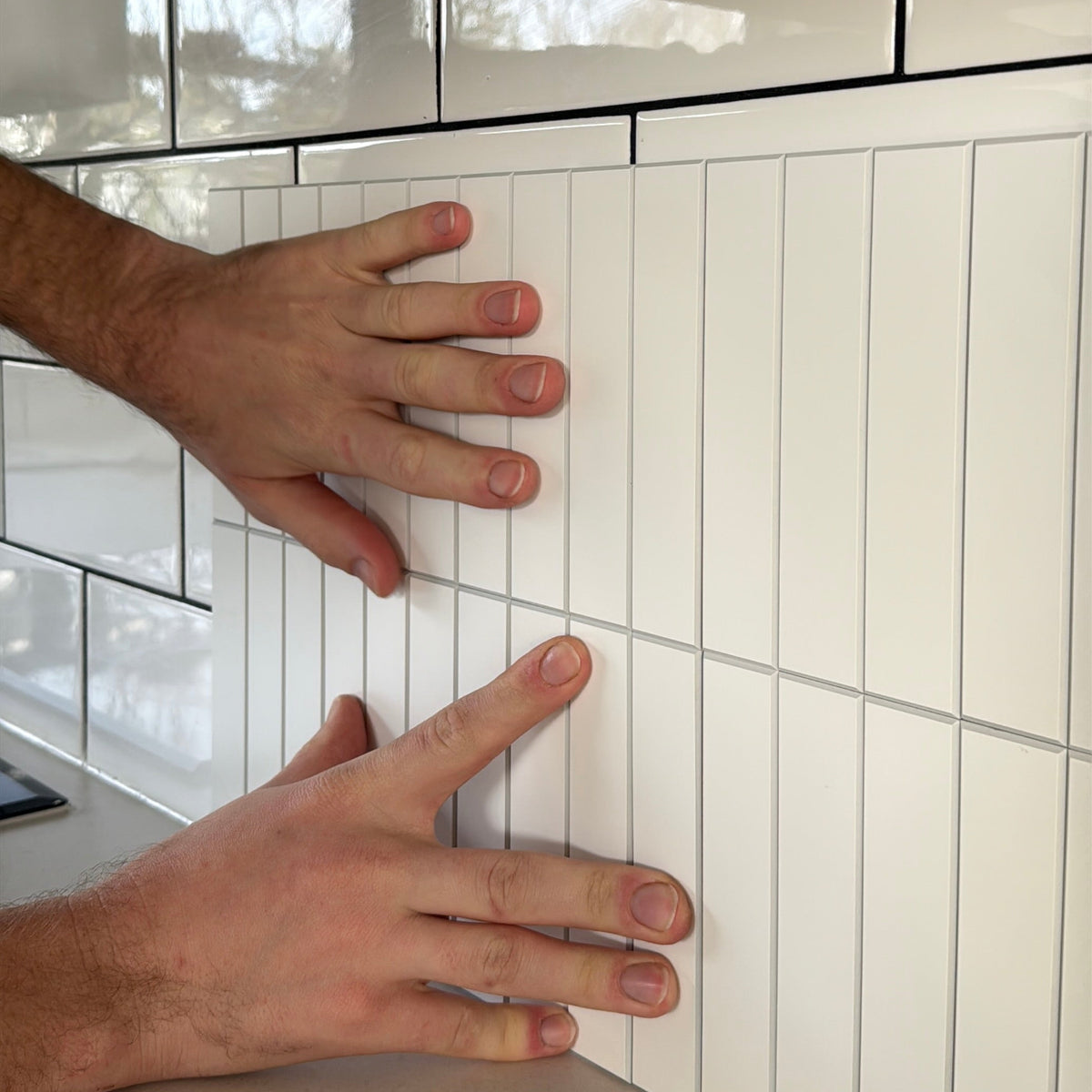 White kit kat wall tiles being installed on a kitchen backsplash, with a person's hands pressing down on the glossy rectangular tiles.