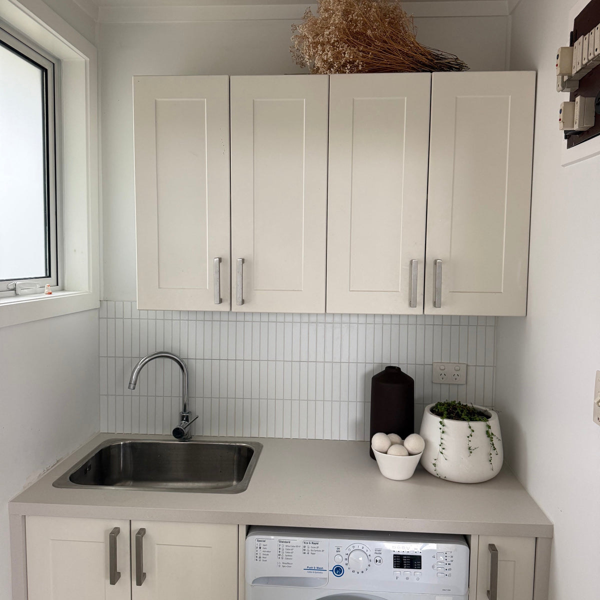 A compact utility room featuring cream-colored shaker-style cabinets, a stainless steel sink, a built-in front-loading washing machine, and a white matte kit kat tile backsplash.