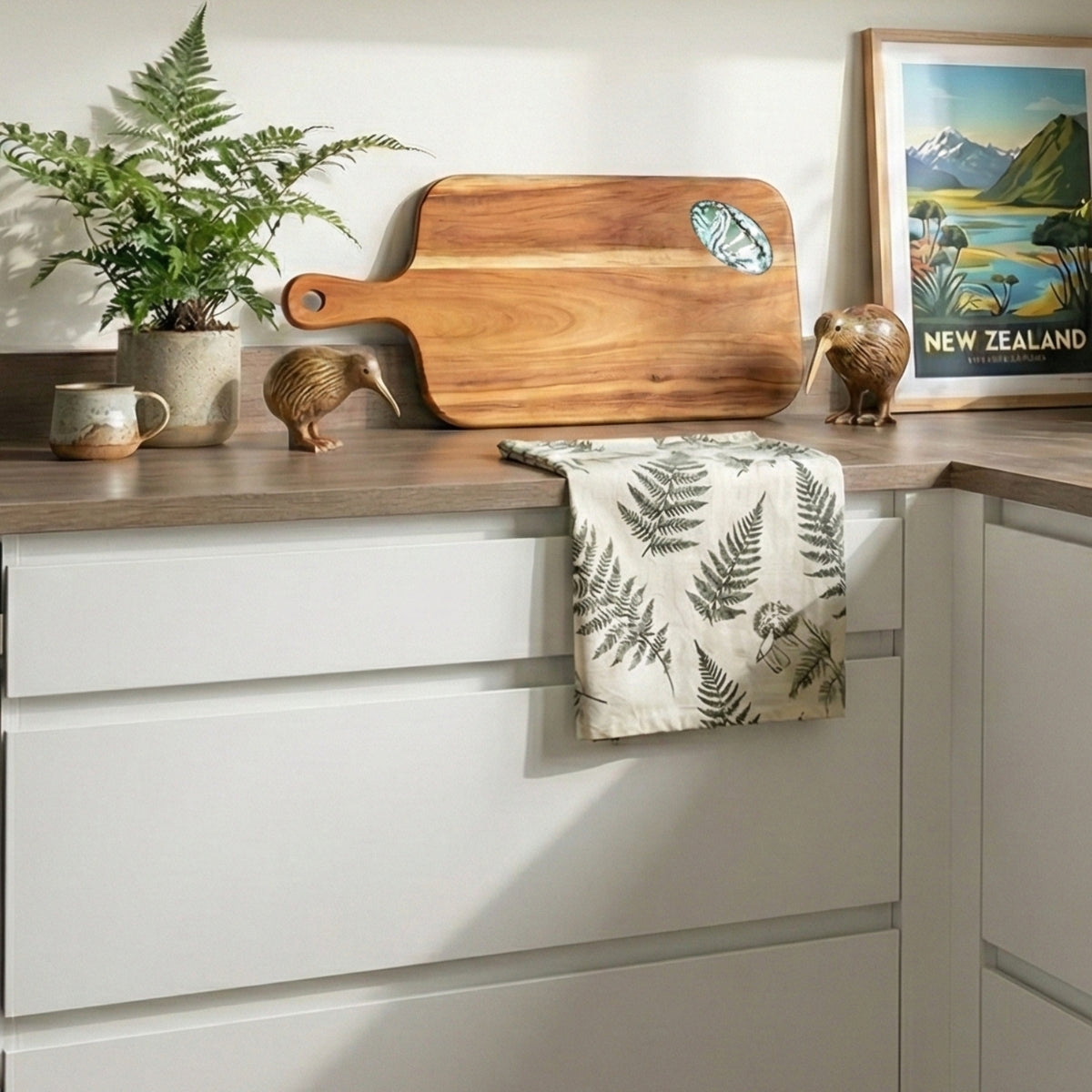 Wooden cutting board on a kitchen counter with a plant, mug, and decorative items.