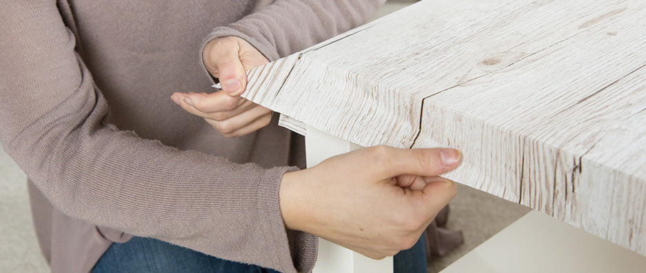 Close up of woman folding down wood grain self-adhesive vinyl over edge on coffee table