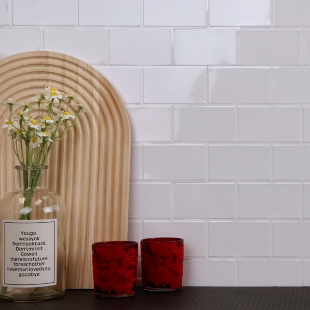 Decorative setup with a glass bottle, red candle holders, and a wooden arch against a white tiled wall.