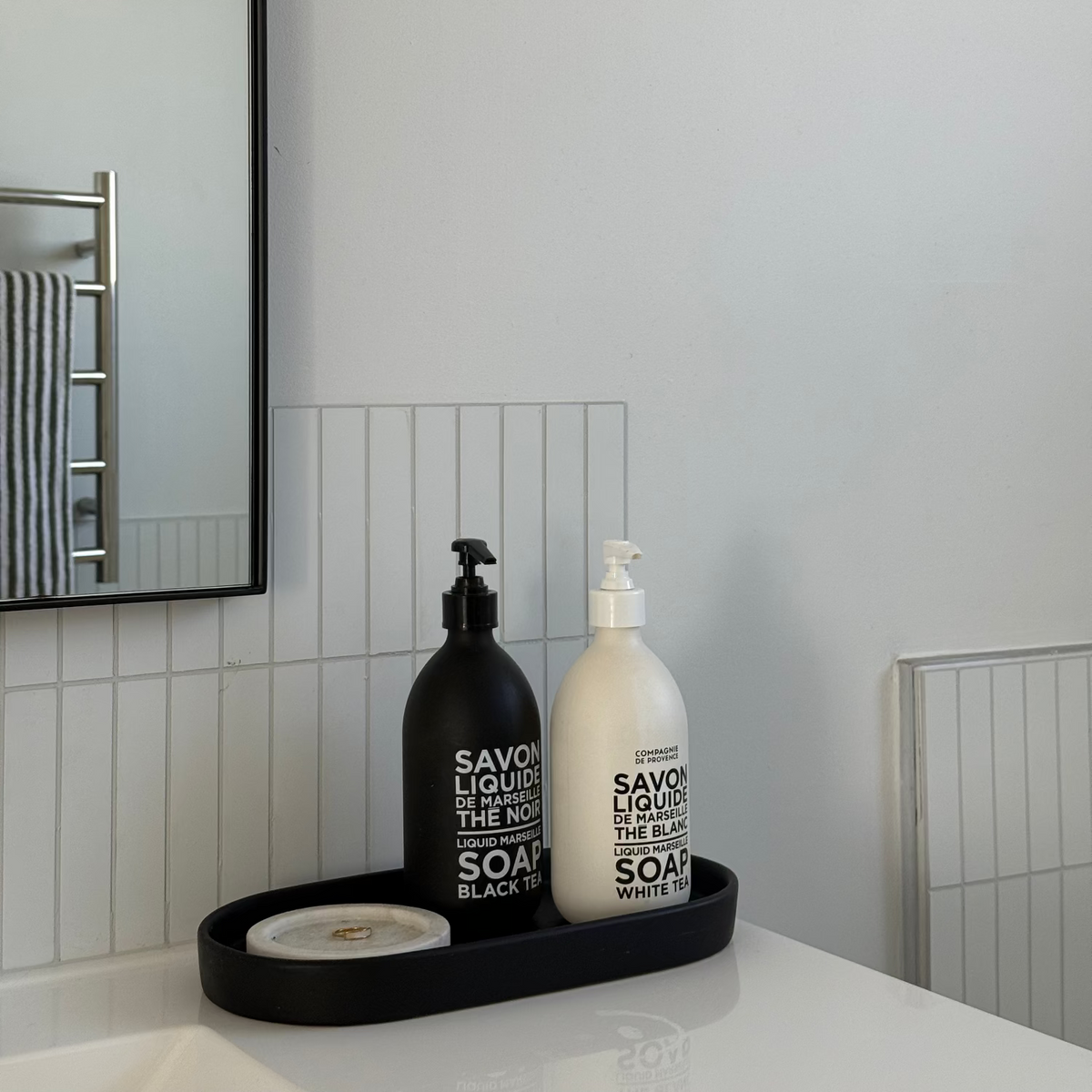 Two bottles of Savon Liquid Hand Soap, one black and one white, displayed on a white countertop in a modern bathroom setting with a mirror and white matte kit kat tiles.