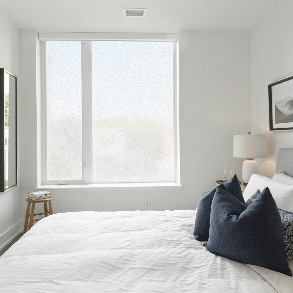 Modern bedroom with a large window film covered window, bed with blue pillows, and lamp.