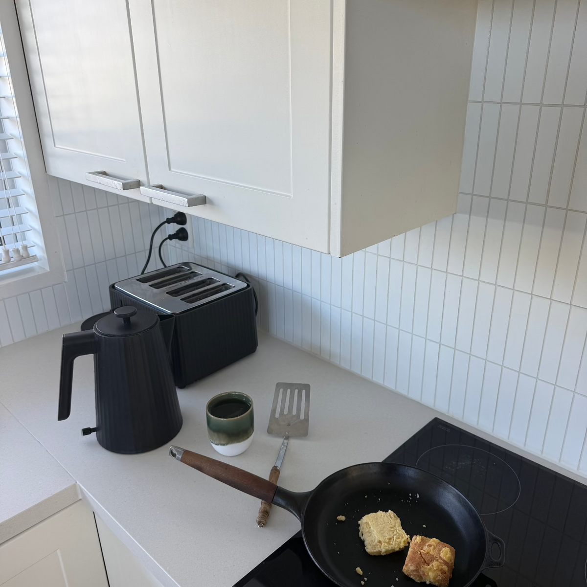 A kitchen countertop scene with a black electric stovetop, a black frying pan cooking two pieces of toast, a black kettle, a toaster, and a small cup on a white tiled backsplash.