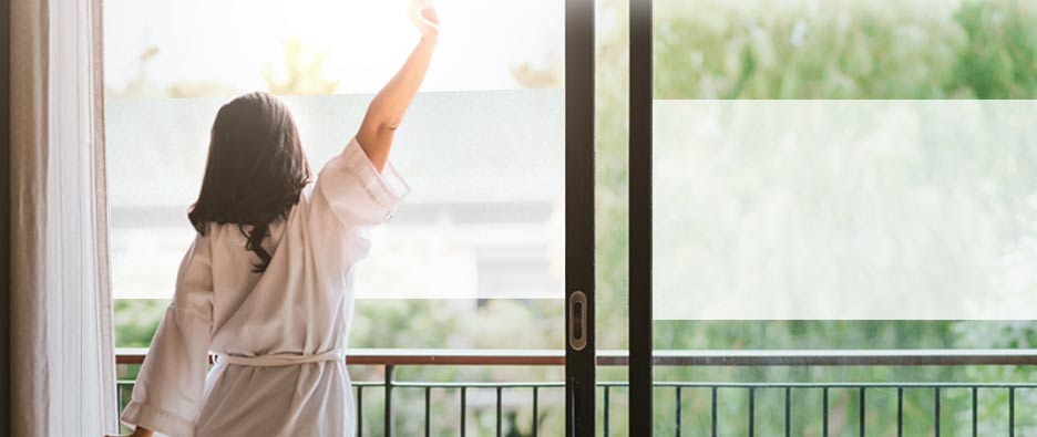 woman in white robe stretching her right arm in front a ranch slider covered with frosted window film