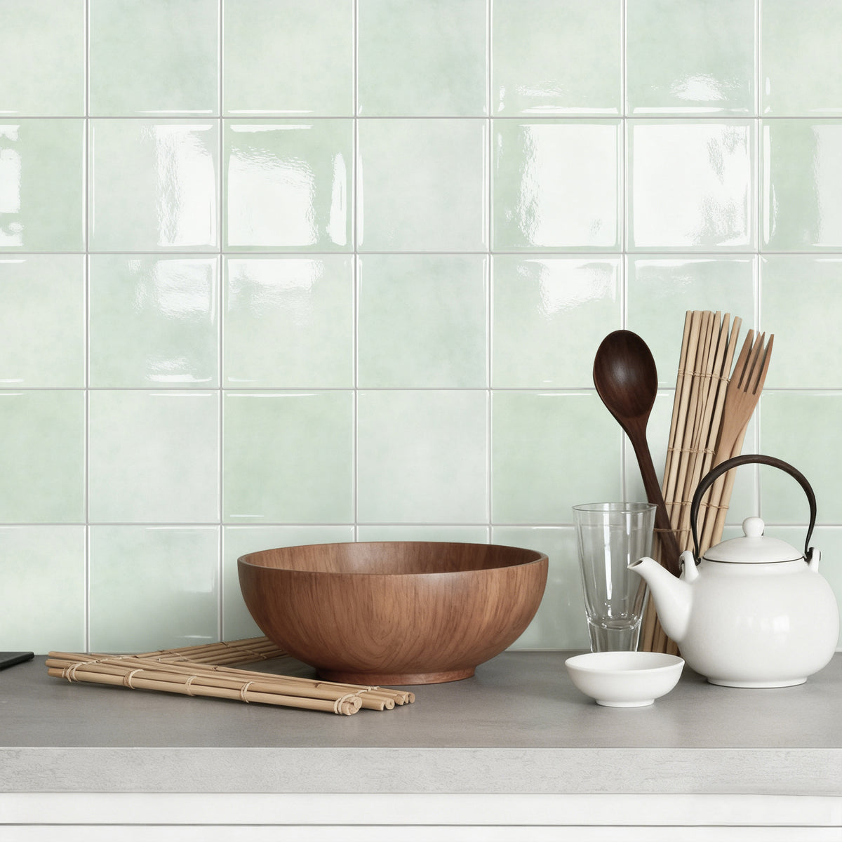 Kitchen counter with wooden bowl, white teapot, and glass against a light green tiled wall.
