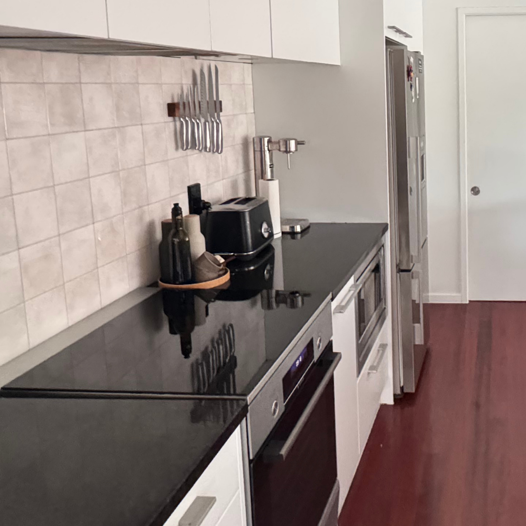 A modern kitchen interior with white cabinetry, black countertops, a tiled backsplash, stainless steel appliances, and reddish-brown wood flooring. A staircase is visible in the background on the right.