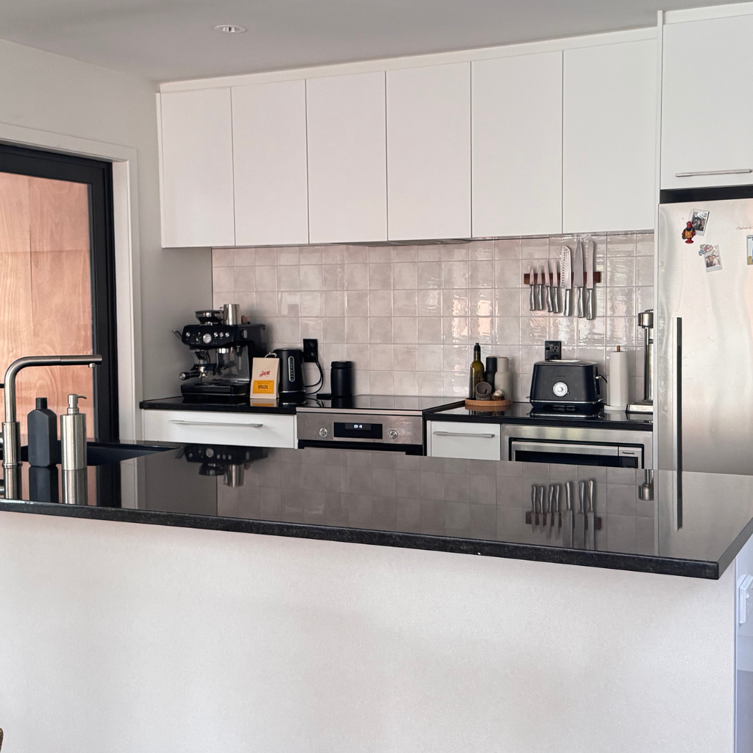 A modern kitchen interior with white upper and lower cabinets, a black countertop, a stainless steel refrigerator, sandy stone tiled backsplash, and small kitchen appliances on the counter.