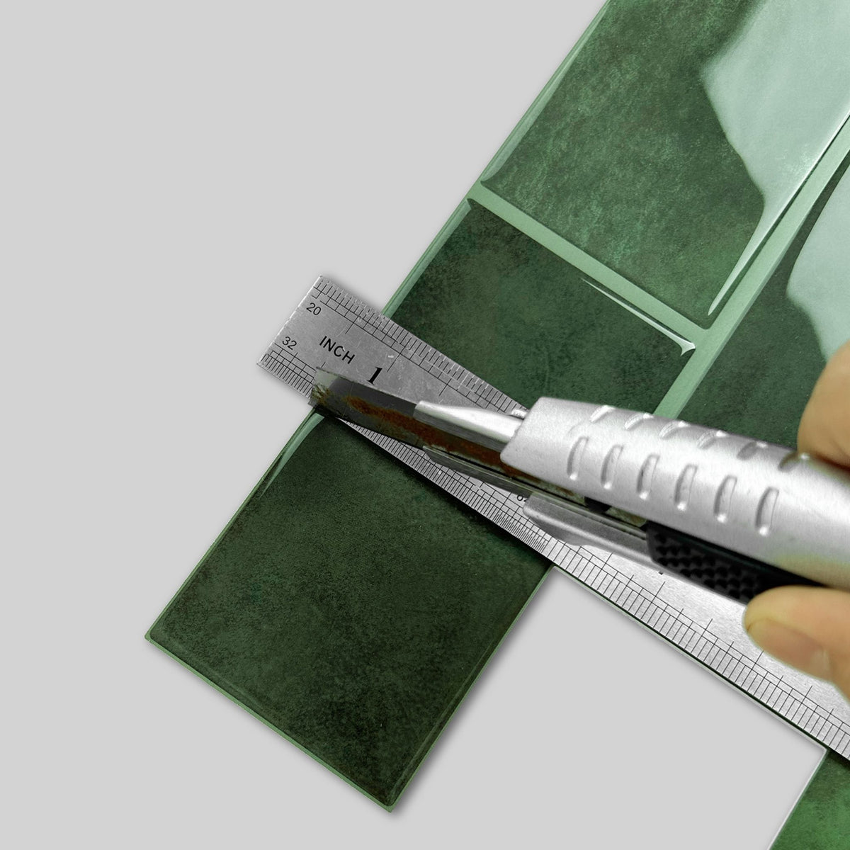 Dark green glossy mosaic tile sheets being measured with a metal ruler and a utility knife, with a hand holding the knife on a light gray background.