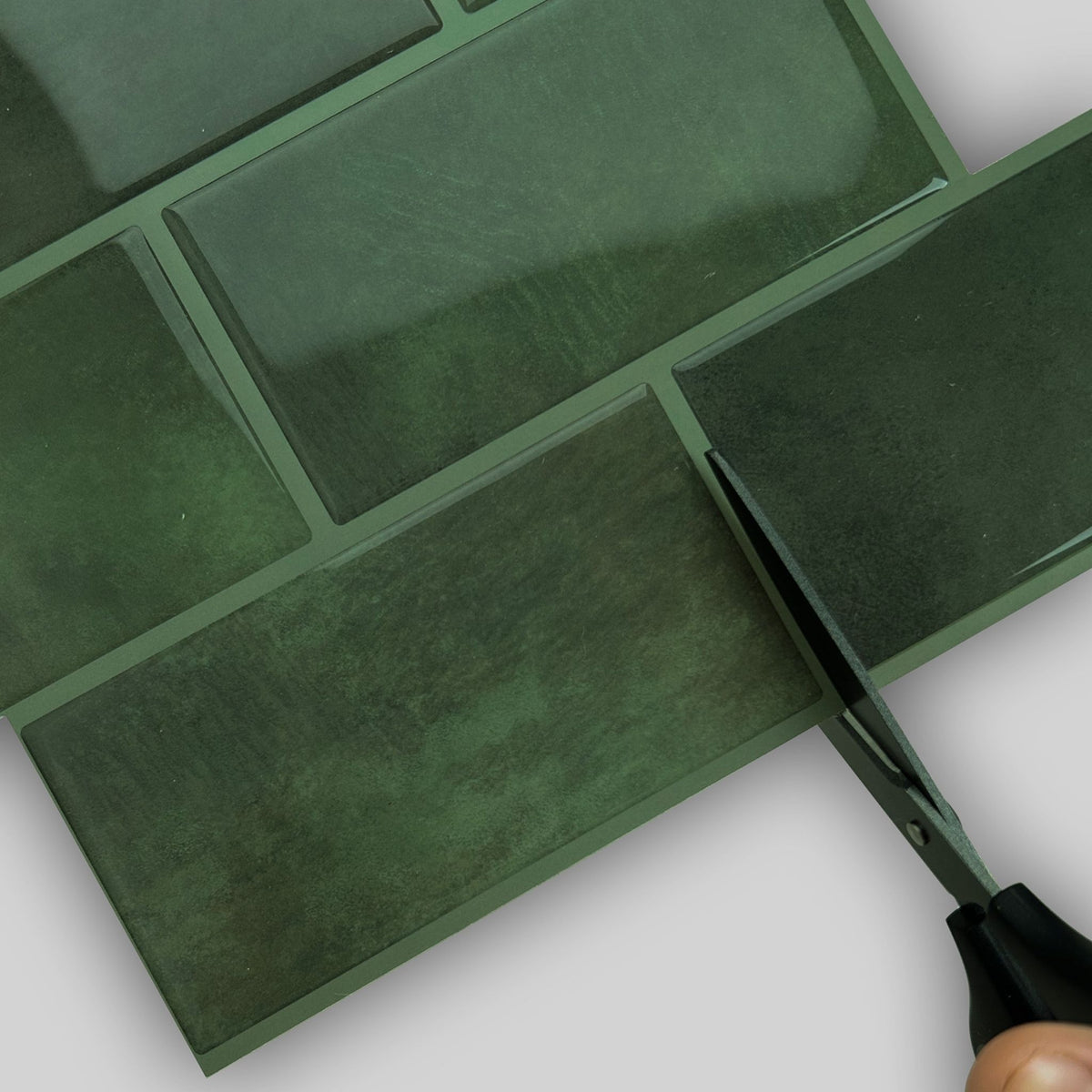Dark green subway tiles with a glossy finish, shown with a hand holding scissors to cut one of the rectangular tiles.