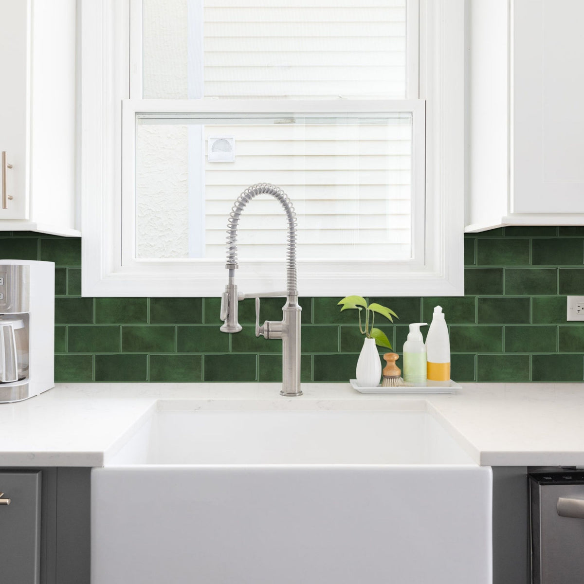 A modern kitchen sink setup featuring a white farmhouse basin, a brushed nickel gooseneck faucet, a white countertop, and a glossy dark green subway tile backsplash behind it.