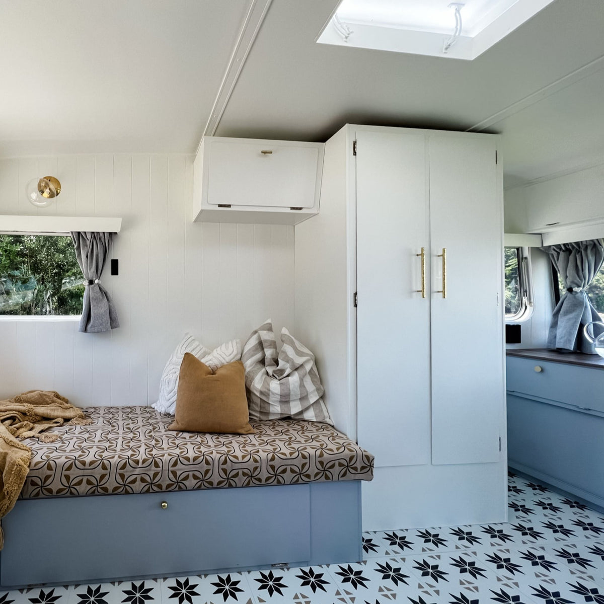 A compact, modern caravan featuring a built-in bed with a brown and beige patterned mattress, a solid brown accent pillow, and a striped throw blanket. White cabinetry with gold handles and a white daybed base are visible, along with a brown and cream star-patterned floor and a window looking out onto green trees.