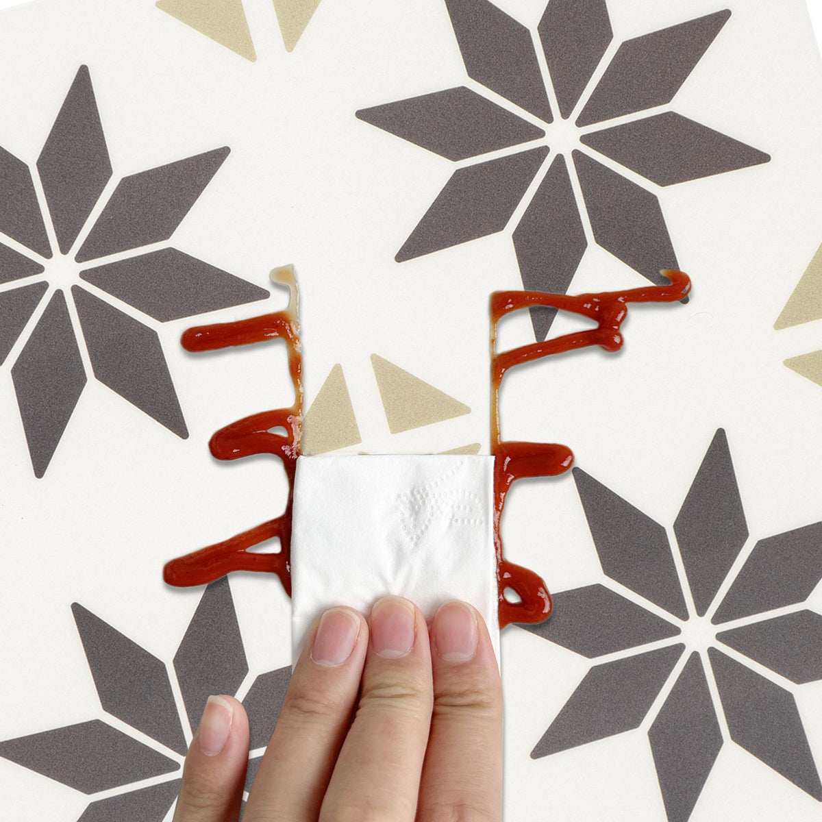 A hand holding a white paper napkin in front of a white surface decorated with gray geometric starburst patterns and brown ketchup drizzles.