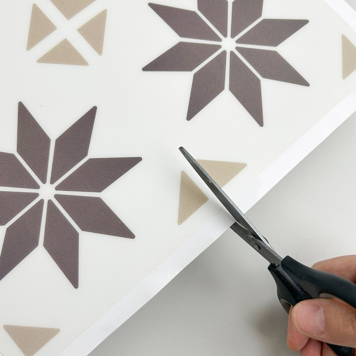 A close-up of a hand holding black-handled scissors and cutting a white sheet printed with geometric star and triangle patterns in taupe and dark brown.