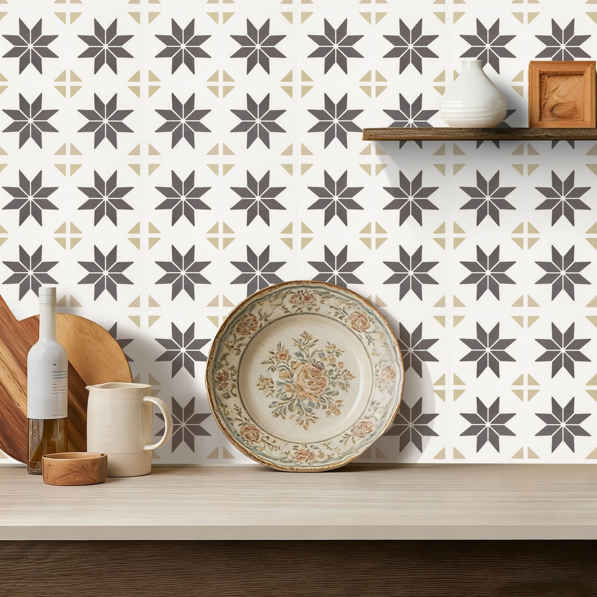 A styled kitchen countertop scene featuring a white wallpaper with a repeating geometric starburst pattern in dark brown and beige. A decorative floral ceramic plate sits on the counter, alongside a mug, a bottle, and a wooden shelf with a white vase and small box in the background.