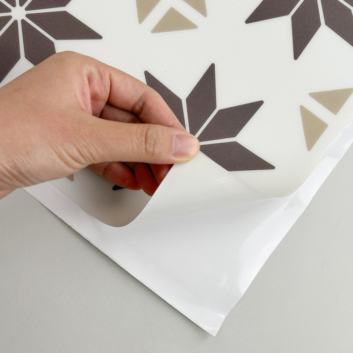 A hand peeling back a clear protective film from a sheet of wall decals featuring a geometric pattern of dark brown and beige starbursts on a white background.