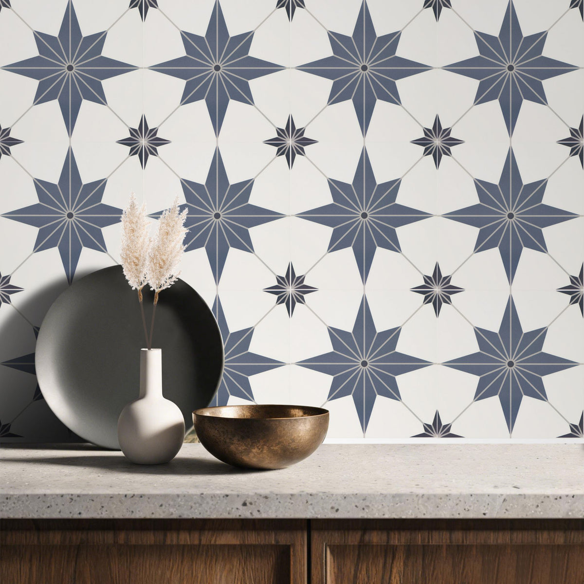 A styled interior scene showing a white wall with repeating navy blue geometric starburst tile pattern, above a stone countertop with a vase, dried grass, and two bowls.