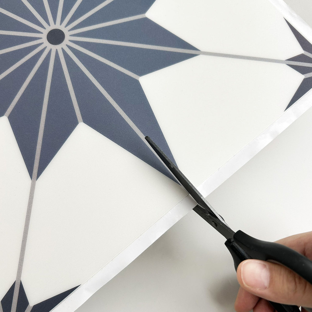 A close-up of a hand holding black-handled scissors and cutting a white sheet with a blue geometric starburst pattern.