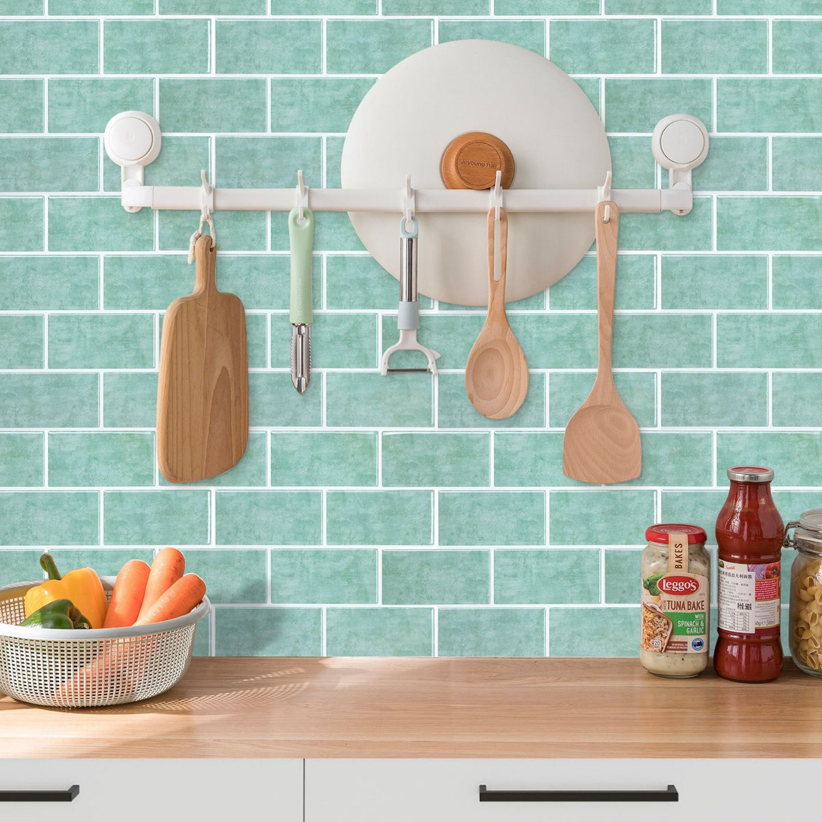 A white wall-mounted kitchen utensil rack with suction cups is shown installed on a mint green subway tile backsplash above a wooden countertop. Hanging from the rack are wooden cutting and serving utensils, including a paddle, spoon, spatula, and peeler, along with a round white holder.