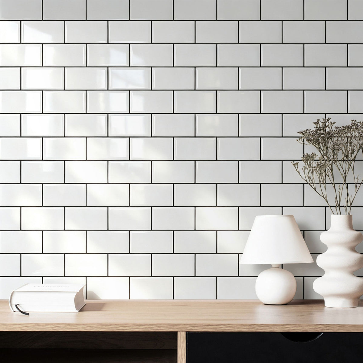 White subway tile wall with a light wood console table, a white table lamp, and a white ceramic vase with dried stems in a bright interior setting.