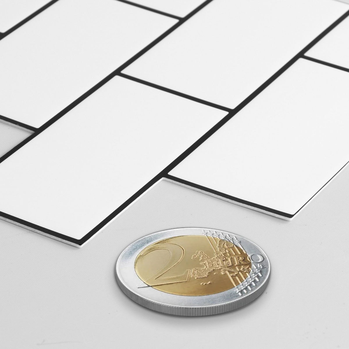 White rectangular wall tiles with black grout lines, shown in a staggered layout on a light gray surface with a two-euro coin for scale.