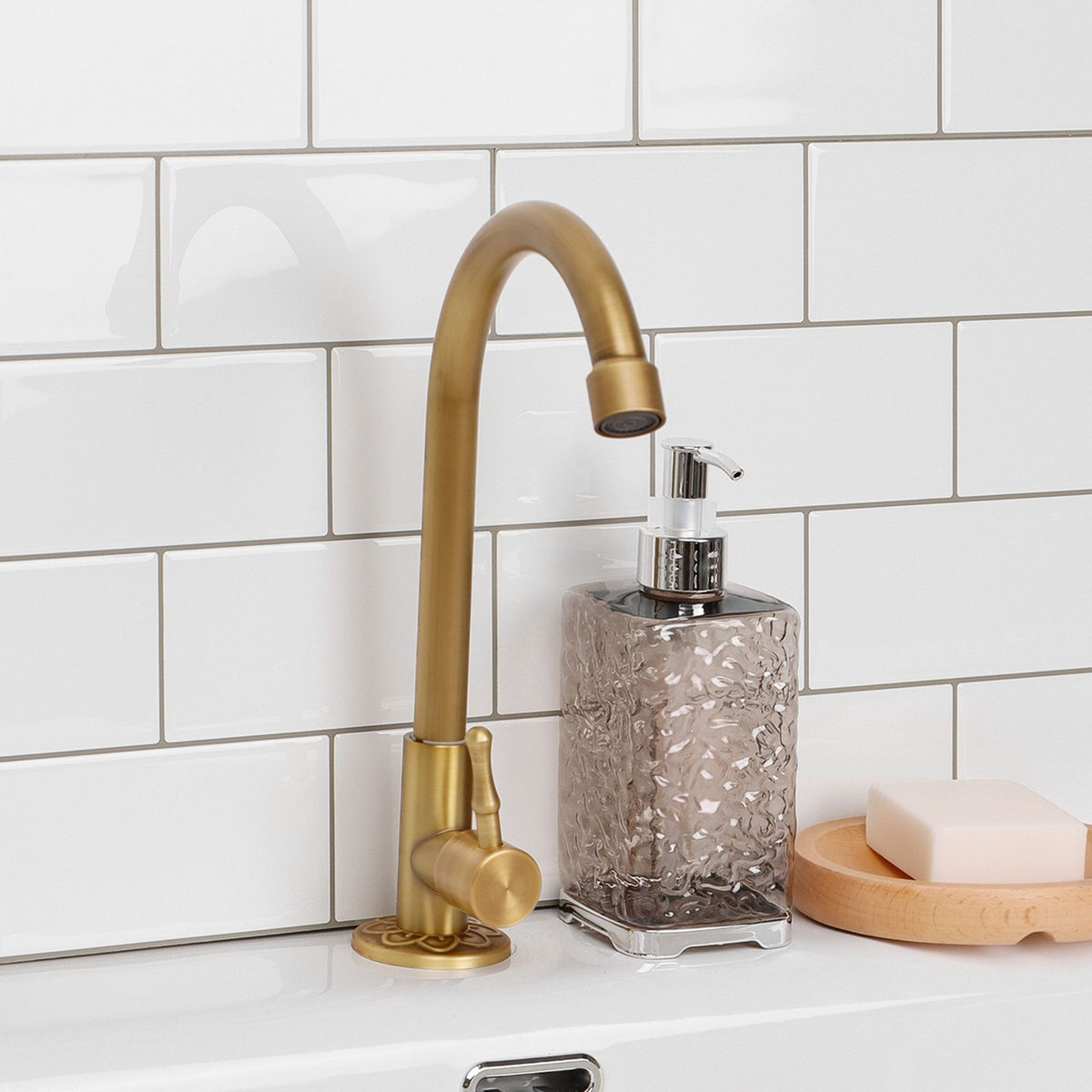 A matte gold kitchen faucet with a curved spout and single lever handle, shown installed on a white sink counter in front of white subway tile. A textured clear soap dispenser and a bar of soap on a wooden soap dish are also visible.