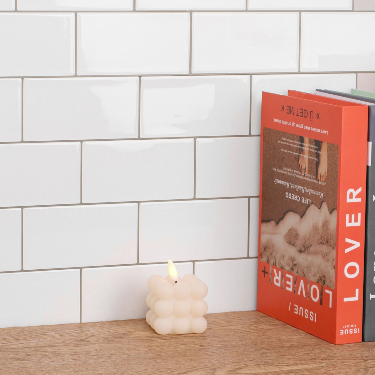 A small cream-colored bubble candle with a lit flame sits on a wooden surface in front of a white subway tile wall, next to a bright orange book titled 'LOVER'.