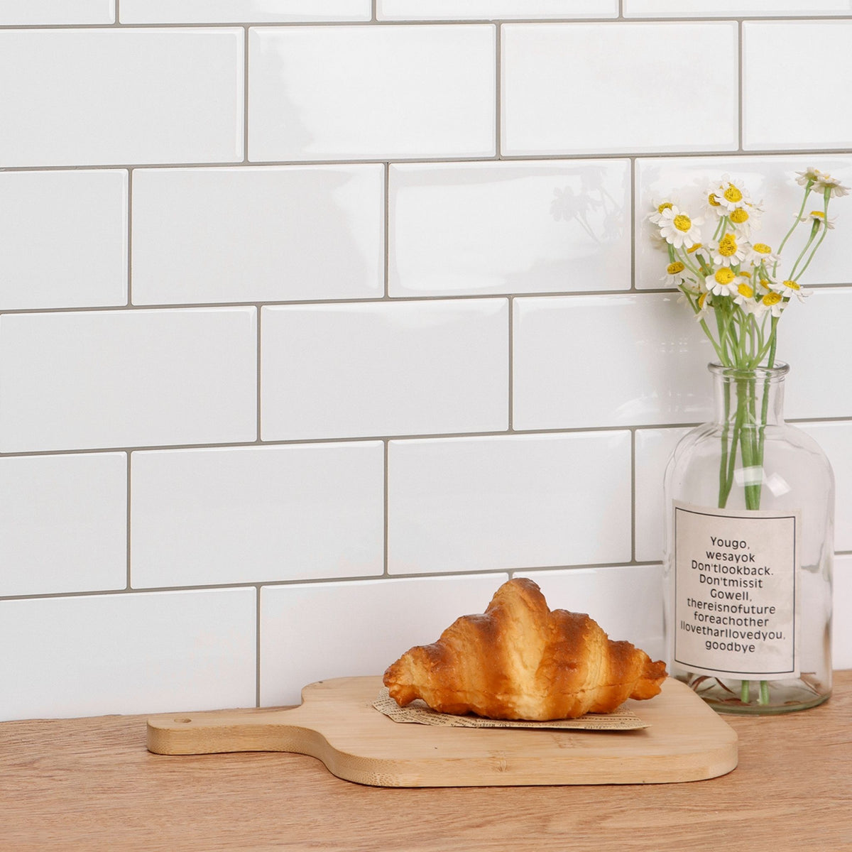 White subway tile backsplash behind a wooden countertop with a croissant on a wooden board and a glass bottle vase with daisies.
