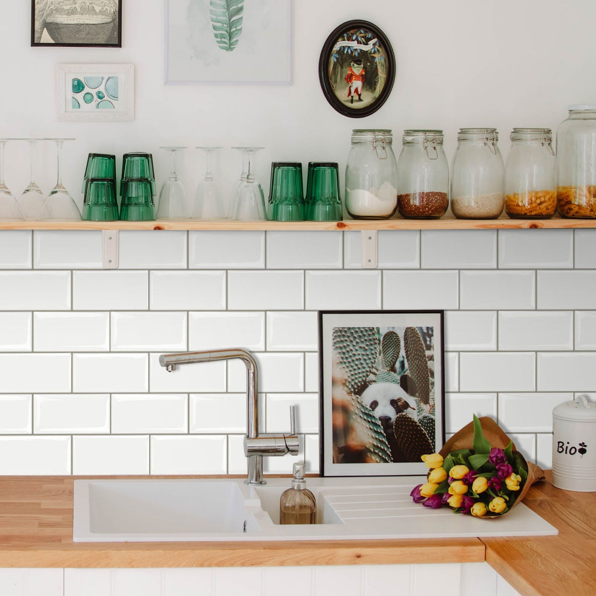 A kitchen scene with a white subway tile backsplash, light wood countertop, a white sink, a chrome faucet, open shelves holding glassware and jars, and framed wall art above.