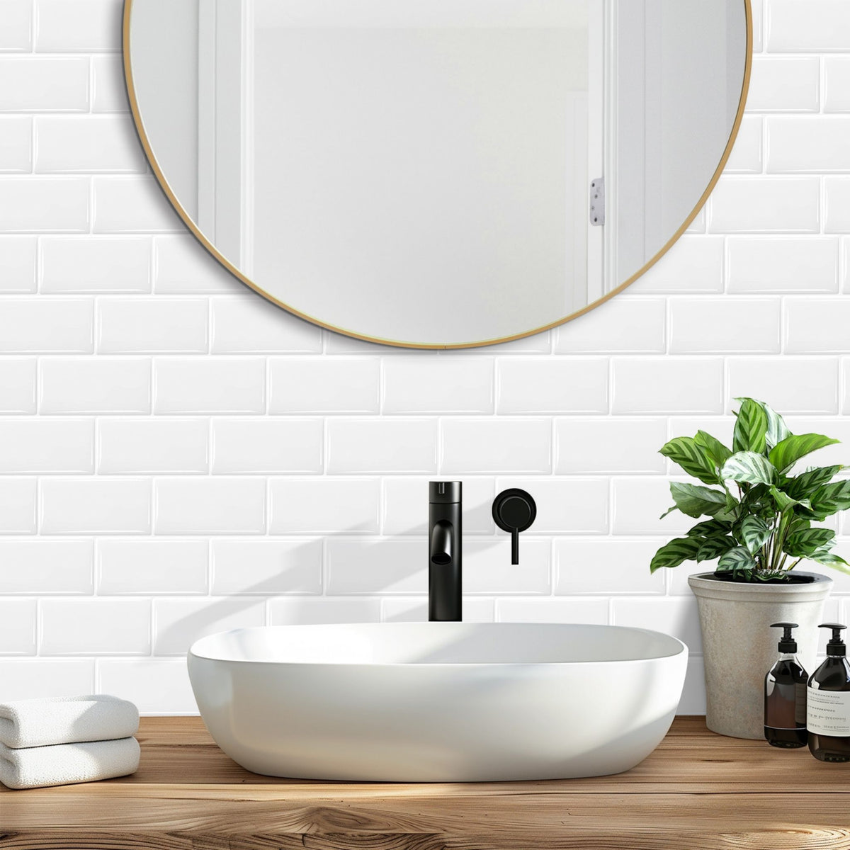 A modern bathroom scene with a white oval vessel sink on a wooden countertop, a matte black faucet, a round wall mirror with a gold frame, white subway tiles, a potted green plant, and small toiletry bottles.
