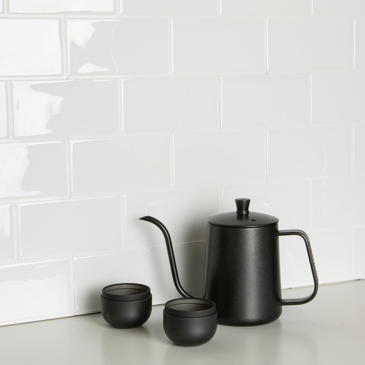 A matte black gooseneck coffee pot with a matching lid and two small black cups, arranged on a light countertop against a white subway tile backsplash.