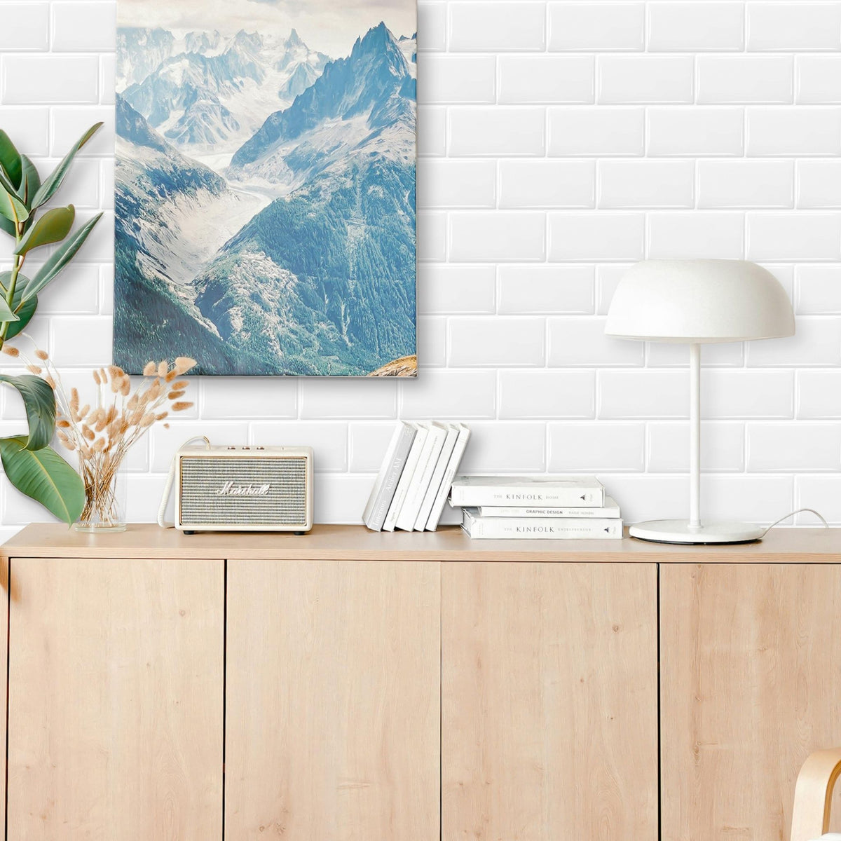 A vertical canvas print of a blue and white mountain landscape is displayed on a white subway tile wall above a light wood cabinet. The scene is styled with a white dome lamp, books, a vintage speaker, and a plant.