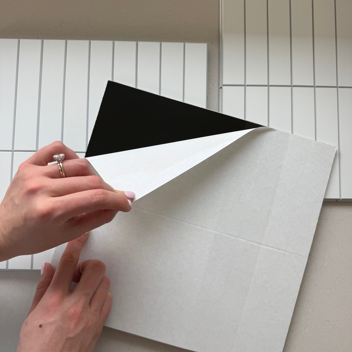 A close-up of a person's hands peeling back a white protective film from a white matte kit kat tile, revealing a black backing layer. In the background, two white rectangular tiles are stacked on a light beige surface.