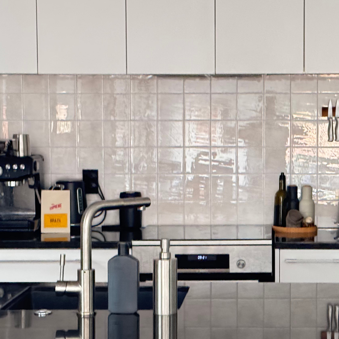 A modern kitchen countertop setup with white cabinets, a glossy sandy tiled backsplash, a black espresso machine, a black toaster, a coffee maker, and a wall-mounted knife rack.