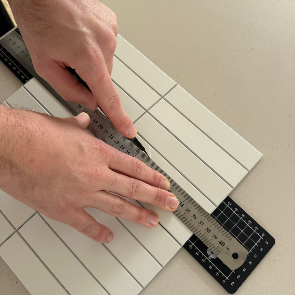 A close-up of a person's hands using a metal ruler and utility knife to measure and cut a white rectangular tile with pre-scored lines, placed on a beige countertop next to a black cutting mat.