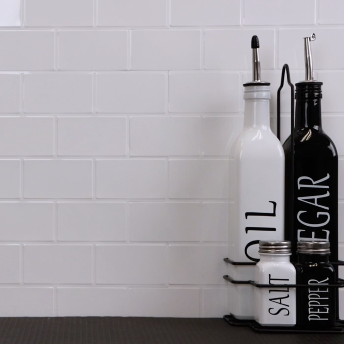 Set of oil, vinegar, salt, and pepper bottles on a kitchen shelf with a white subway tiled wall background.