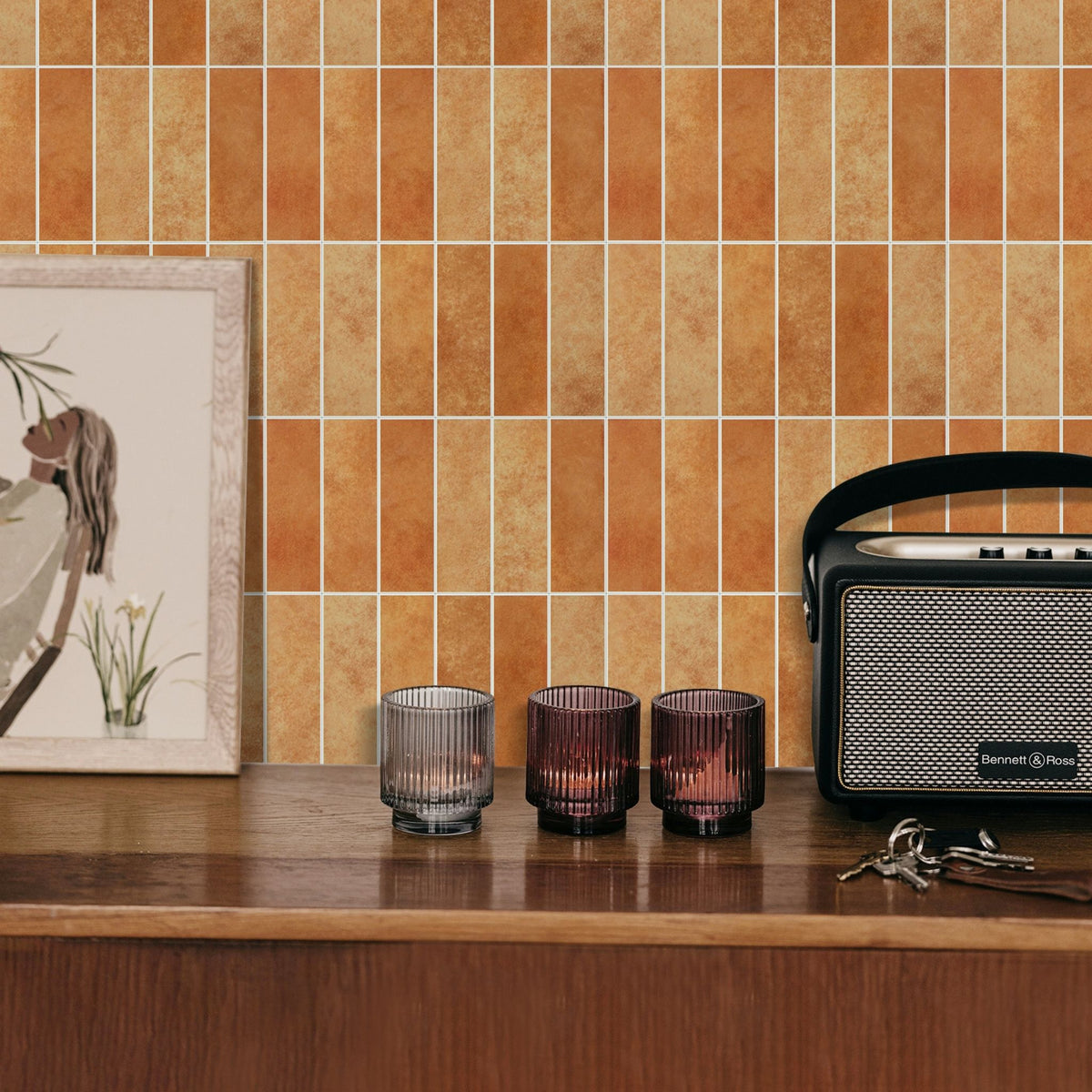 Orange and brown rectangular composite wall tiles with a white grout line, shown installed on a kitchen backsplash behind a wooden counter with three ribbed glass candle holders, a framed print, and a vintage radio.