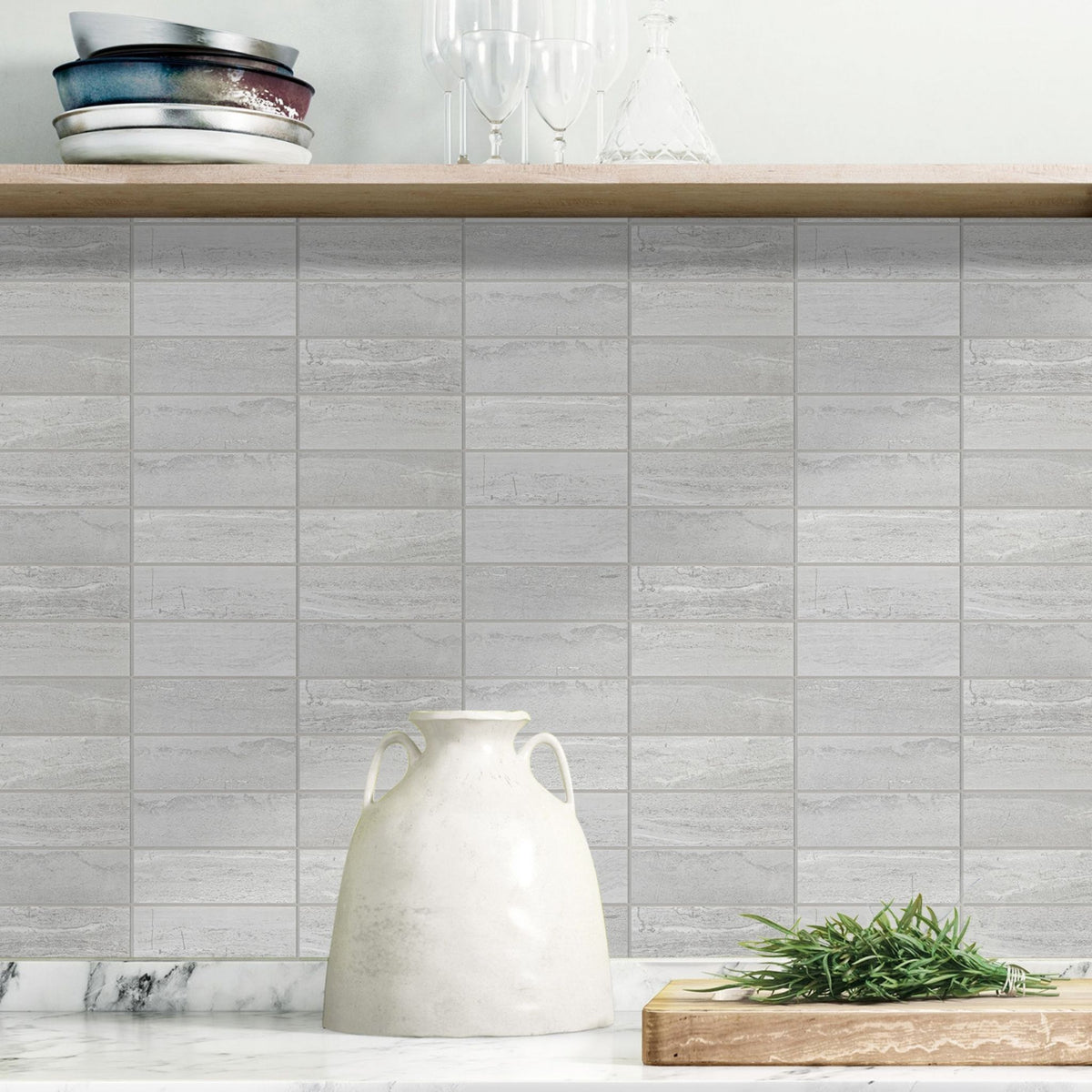 Light gey concrete-look rectangular subway tiles installed on a kitchen backsplash, with a marble countertop below, a white ceramic vase in the foreground, and a wooden shelf with dishes and glassware above.