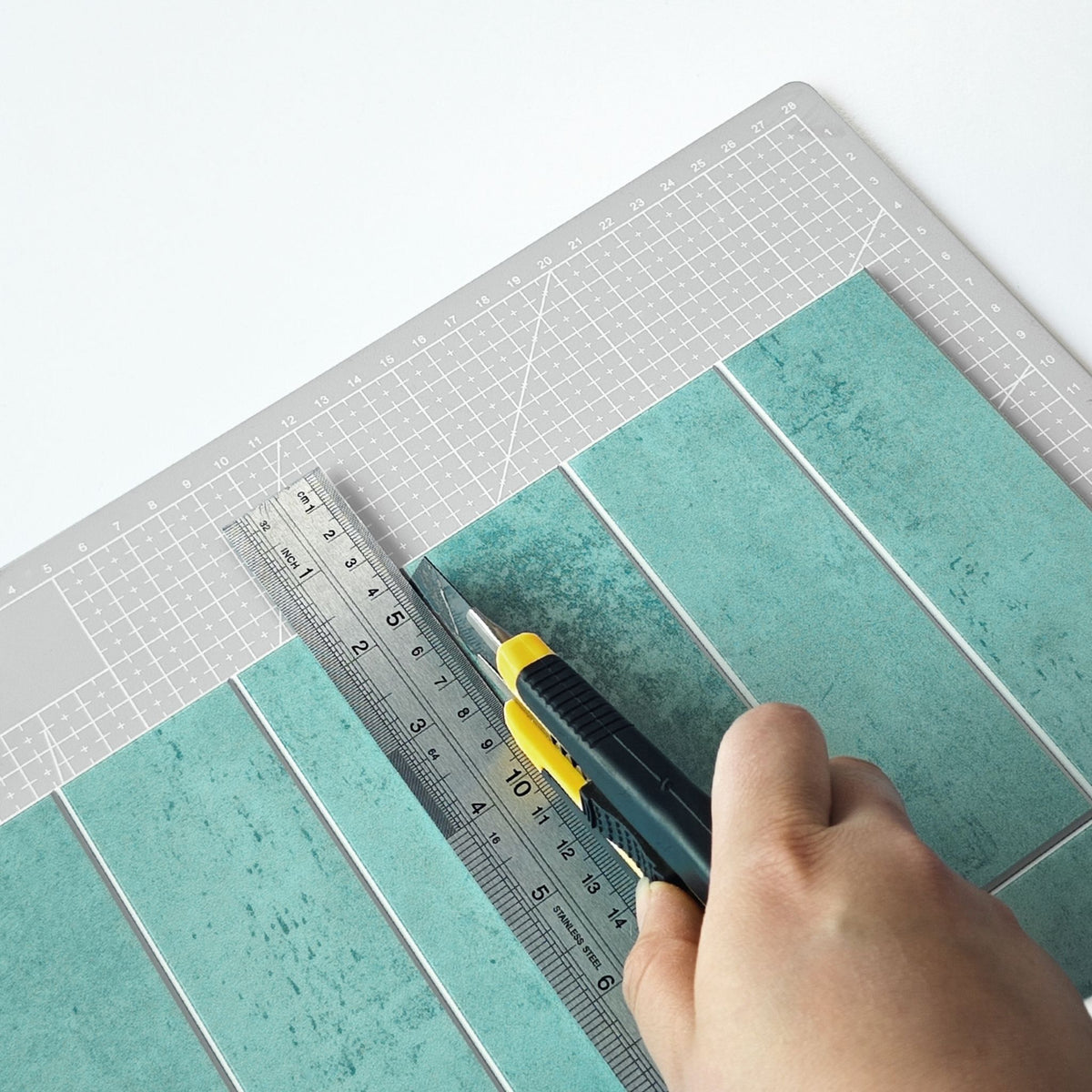 A hand uses a yellow and black utility knife with a metallic ruler to cut teal textured rectangular tiles on a grey gridded cutting mat.